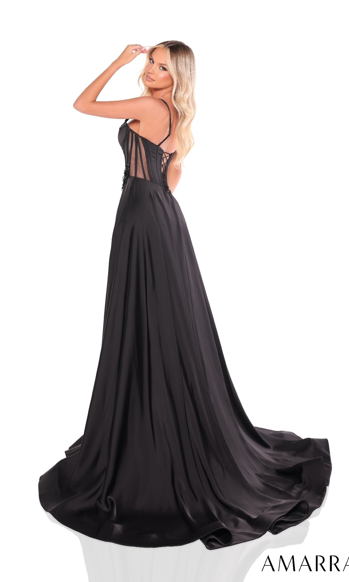 Woman wears the Amarra 88214 long prom dress with a black satin corset bodice, sheer back, and high leg slit, standing on a white background and glancing over her shoulder.